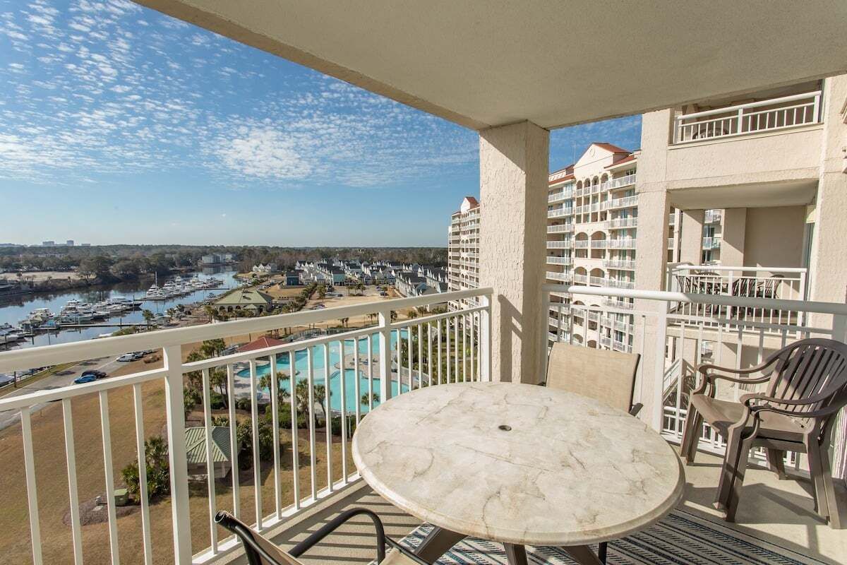 A balcony with a round table and chairs, overlooking a marina with boats and a swimming pool.