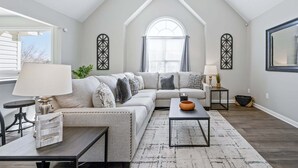 A loft-style living room with a high ceiling, a large window, a sectional sofa, a coffee table, and a side table with a lamp. - 126 Manning Mill Adairsville (Adairsville)