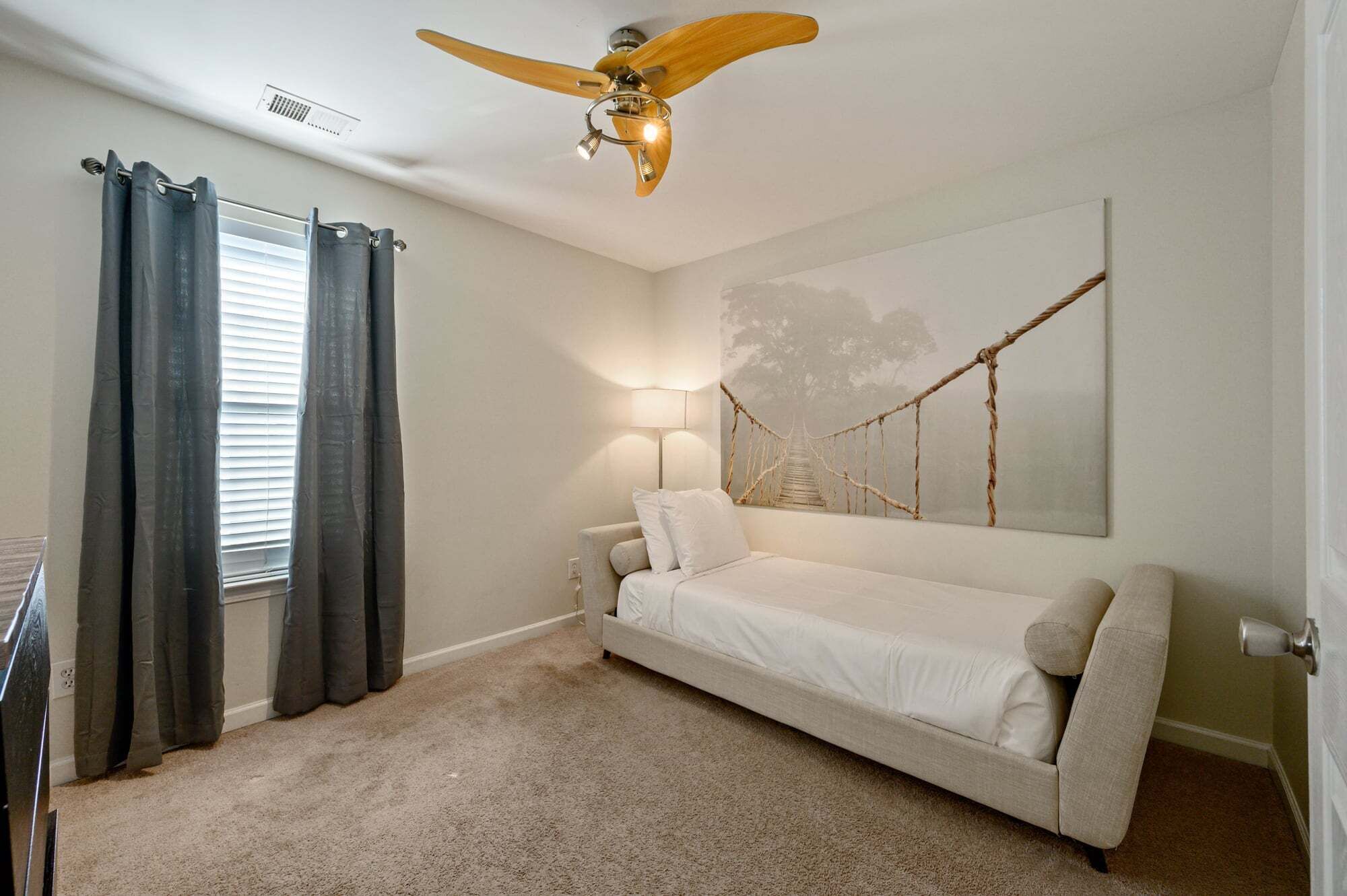 A bedroom with a bed, a ceiling fan, a lamp, a window with curtains, and a wall art piece.