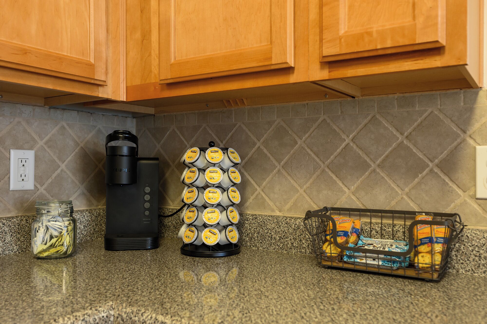 A kitchen with wooden cabinets, a coffee maker, a mug holder, a jar, and a basket with bread.