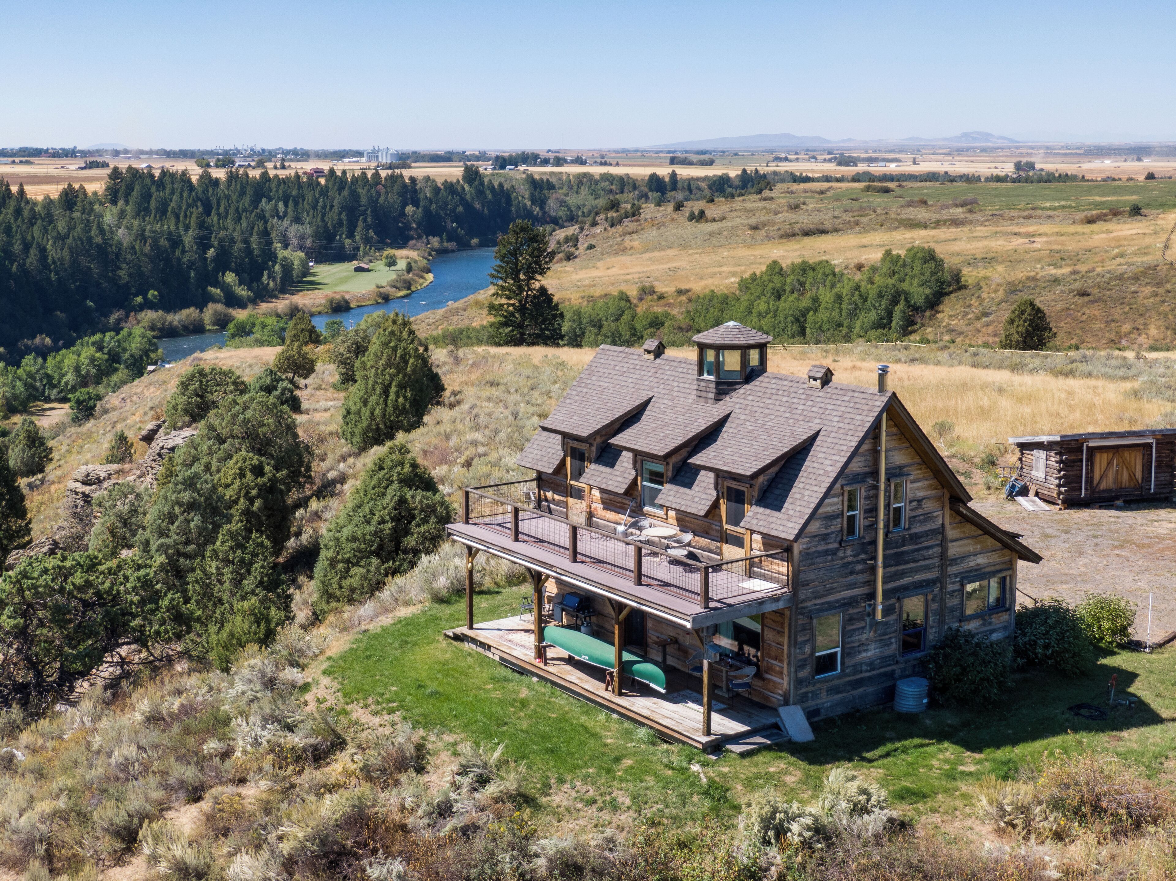 A log cabin with a deck, overlooking a river and forested landscape.