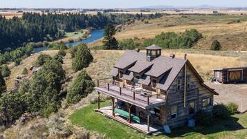A log cabin with a deck, overlooking a river and forested landscape.