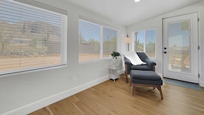 A spacious room with large windows, a grey armchair, a small table with a lamp, and a door with a glass panel. - Boulder Apache Junction (Apache Junction)