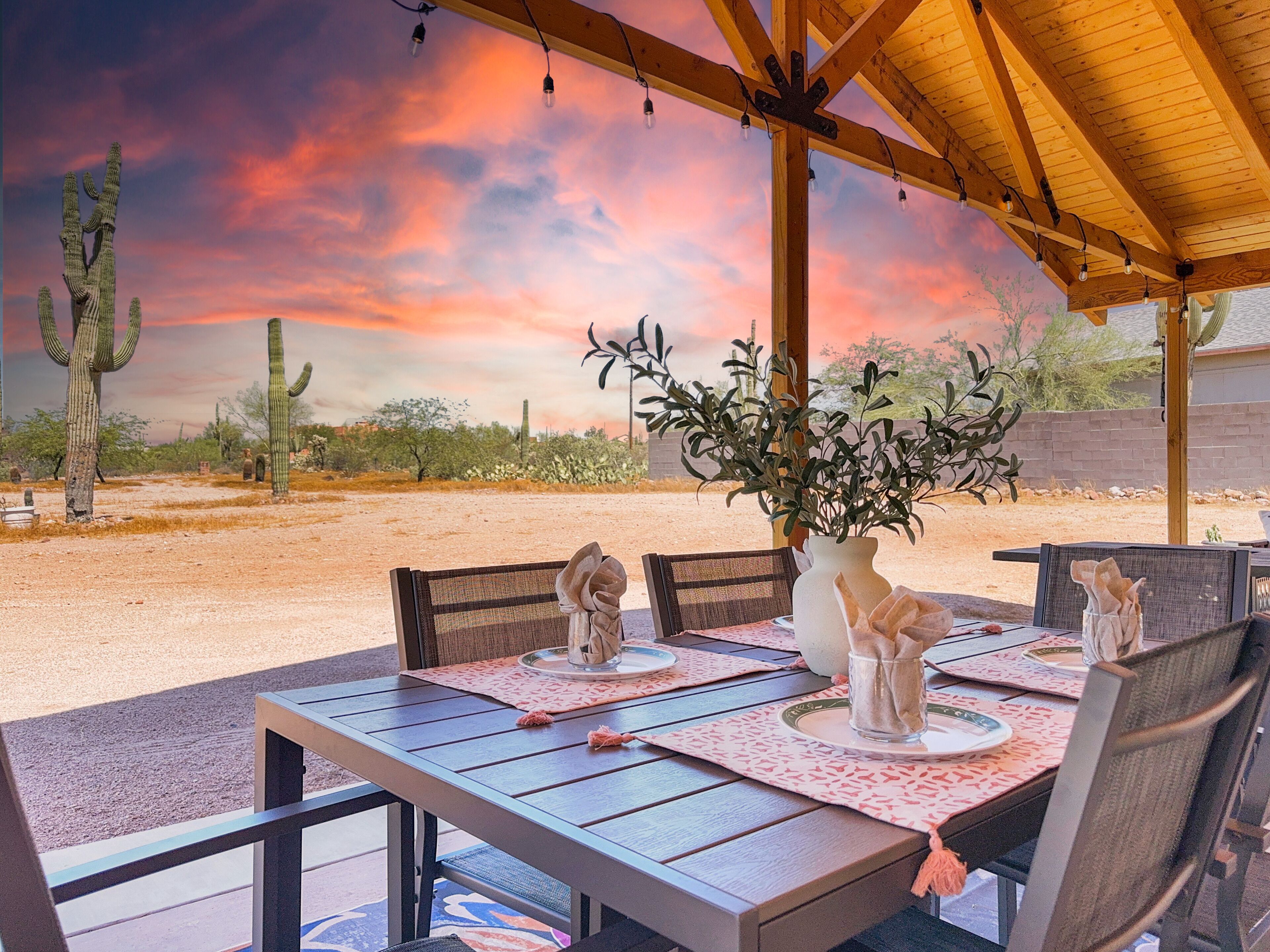An outdoor dining area with a table set for four, surrounded by chairs, under a wooden pergola with string lights.