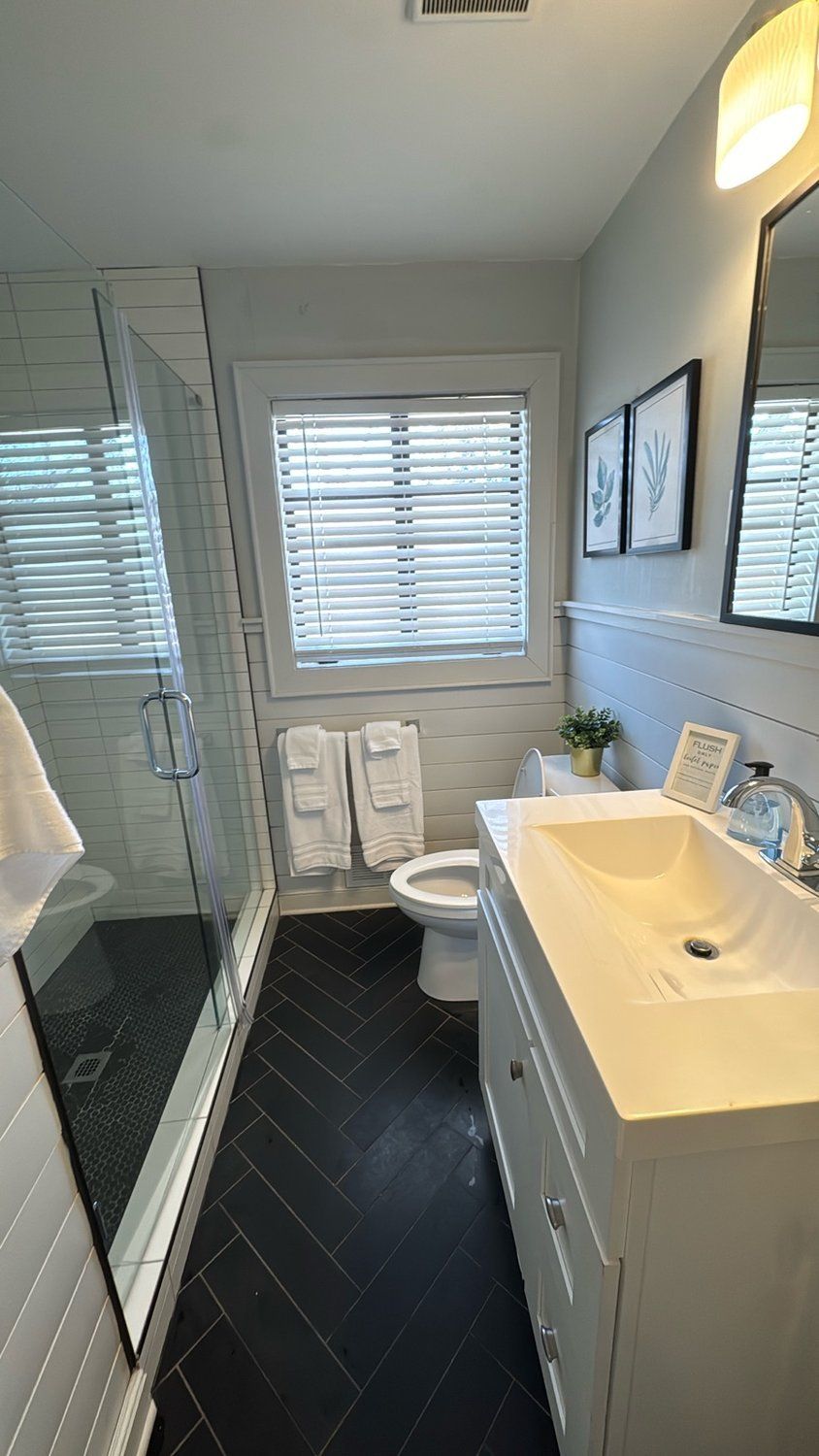 A modern bathroom with a glass shower enclosure, a toilet, a sink with a mirror, and a window with blinds.