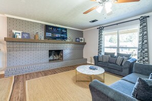 Ein Wohnzimmer mit Kamin aus Ziegeln, Deckenventilator, grauem Sofa und rundem Couchtisch.