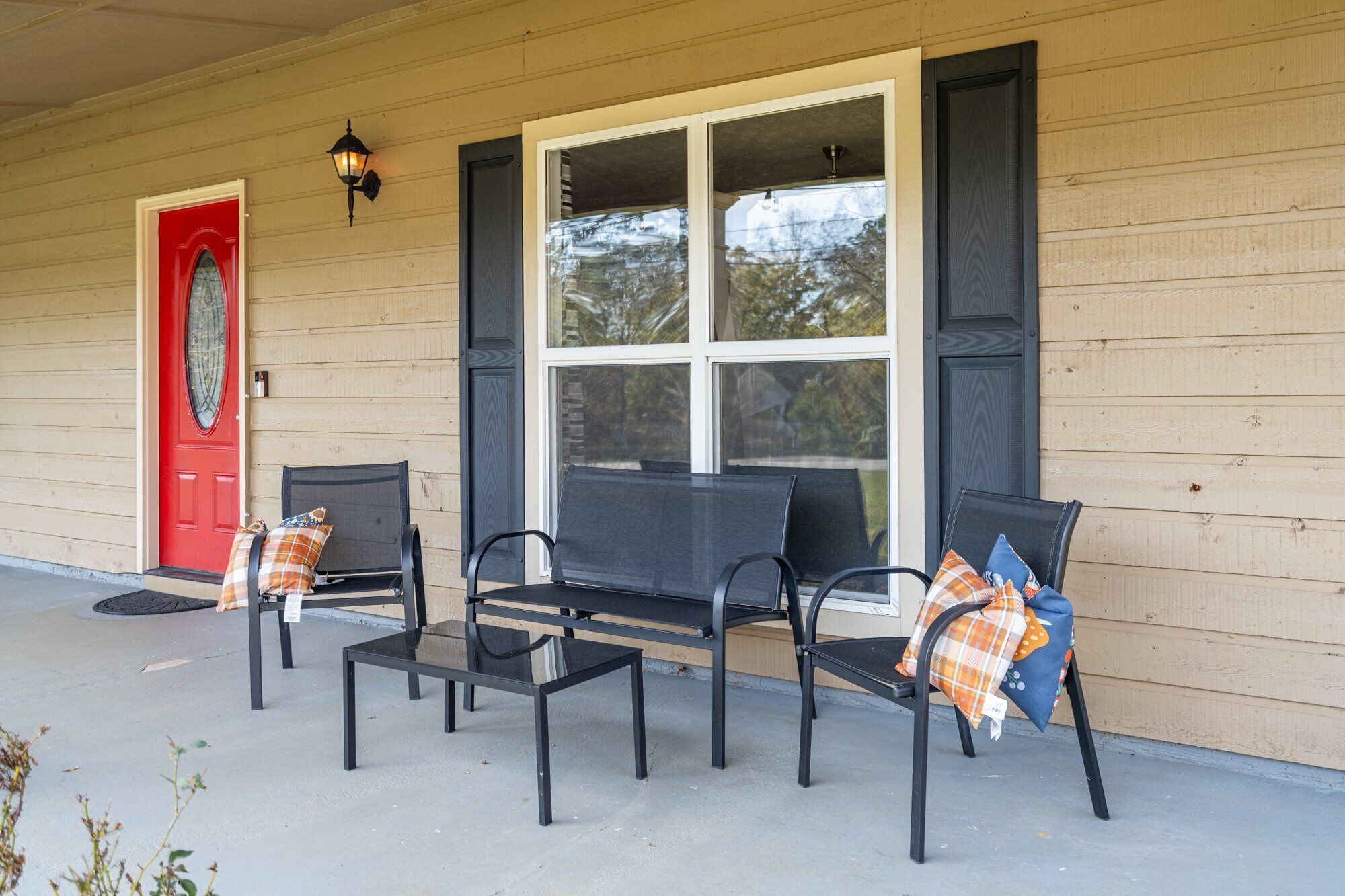 A porch with two black metal chairs and a small table, a red door, and a large window with white frames.
