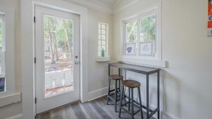 A hallway with a door leading to a covered outdoor area, a window with a view of trees, and a small table with two stools. - Five Points Mill House (Athens)