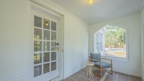 A covered porch with a white brick wall, a glass door, a small round table with a chair, and a window with a view of greenery. - Five Points Mill House (Athens)