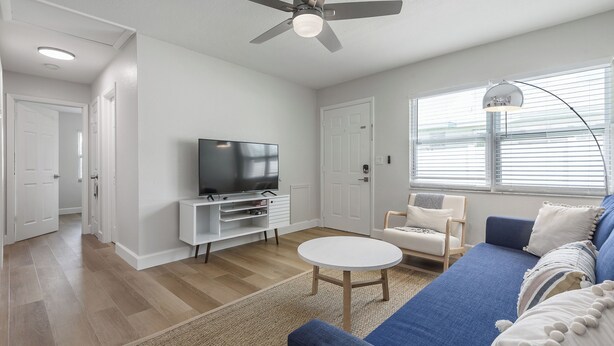 Ein modernes Wohnzimmer mit Flachbildfernseher, Deckenventilator, rundem Couchtisch, blauem Sofa und einem Fenster mit Jalousien.