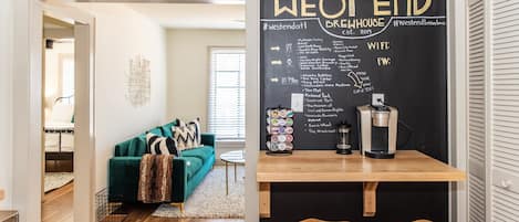 Uma sala de estar aconchegante com um sofá verde-água, uma máquina de café e um quadro negro com lista de atrações e serviços locais.