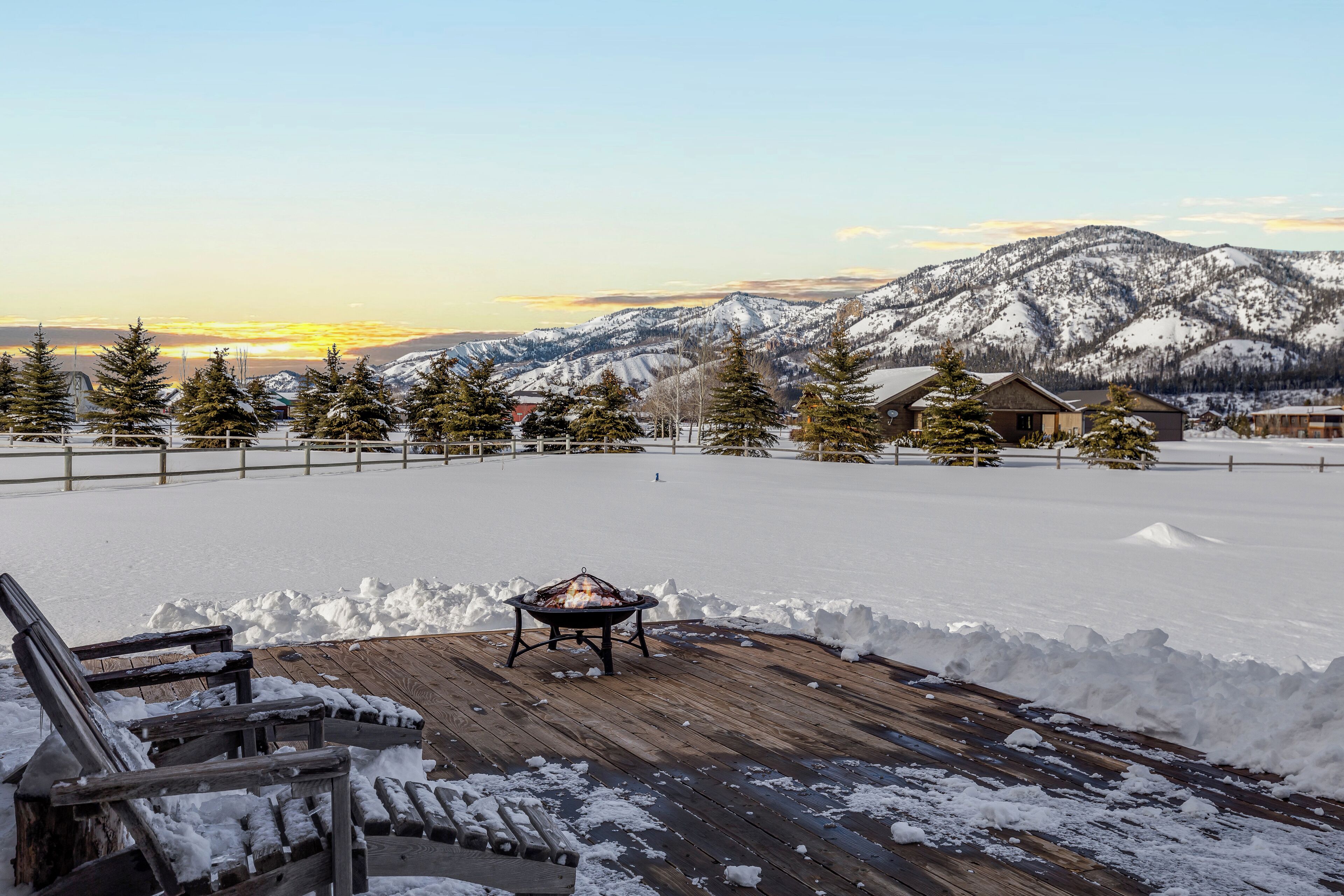 Una terraza de madera con un brasero, rodeada de nieve y montañas al fondo.