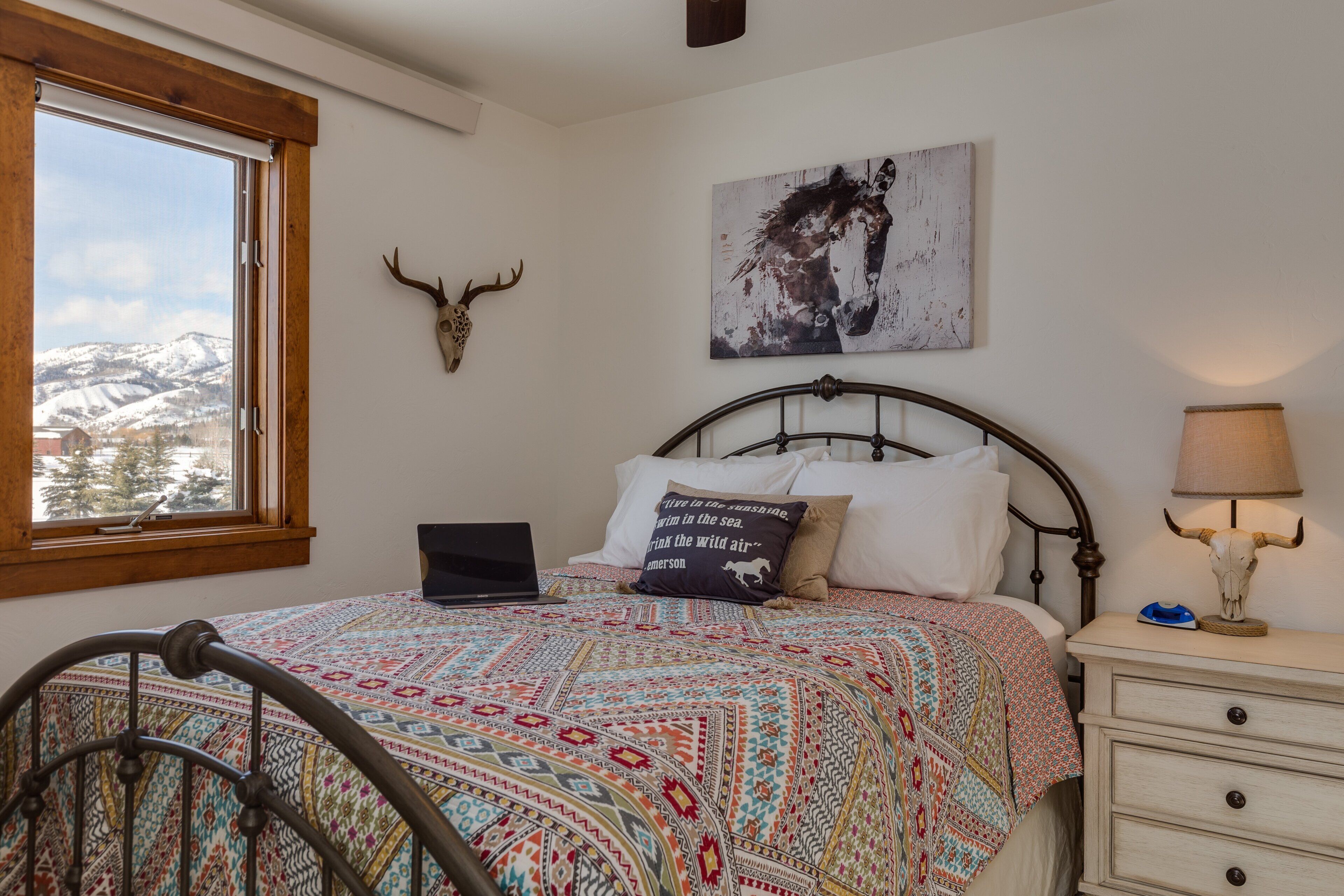 A bedroom with a bed, a laptop, a lamp, a wall-mounted deer head, and a painting.