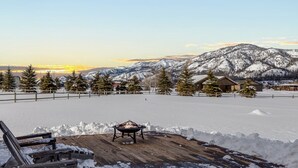 Eine Holzterrasse mit einem Feuerkorb, umgeben von schneebedecktem Boden und Bergen im Hintergrund.