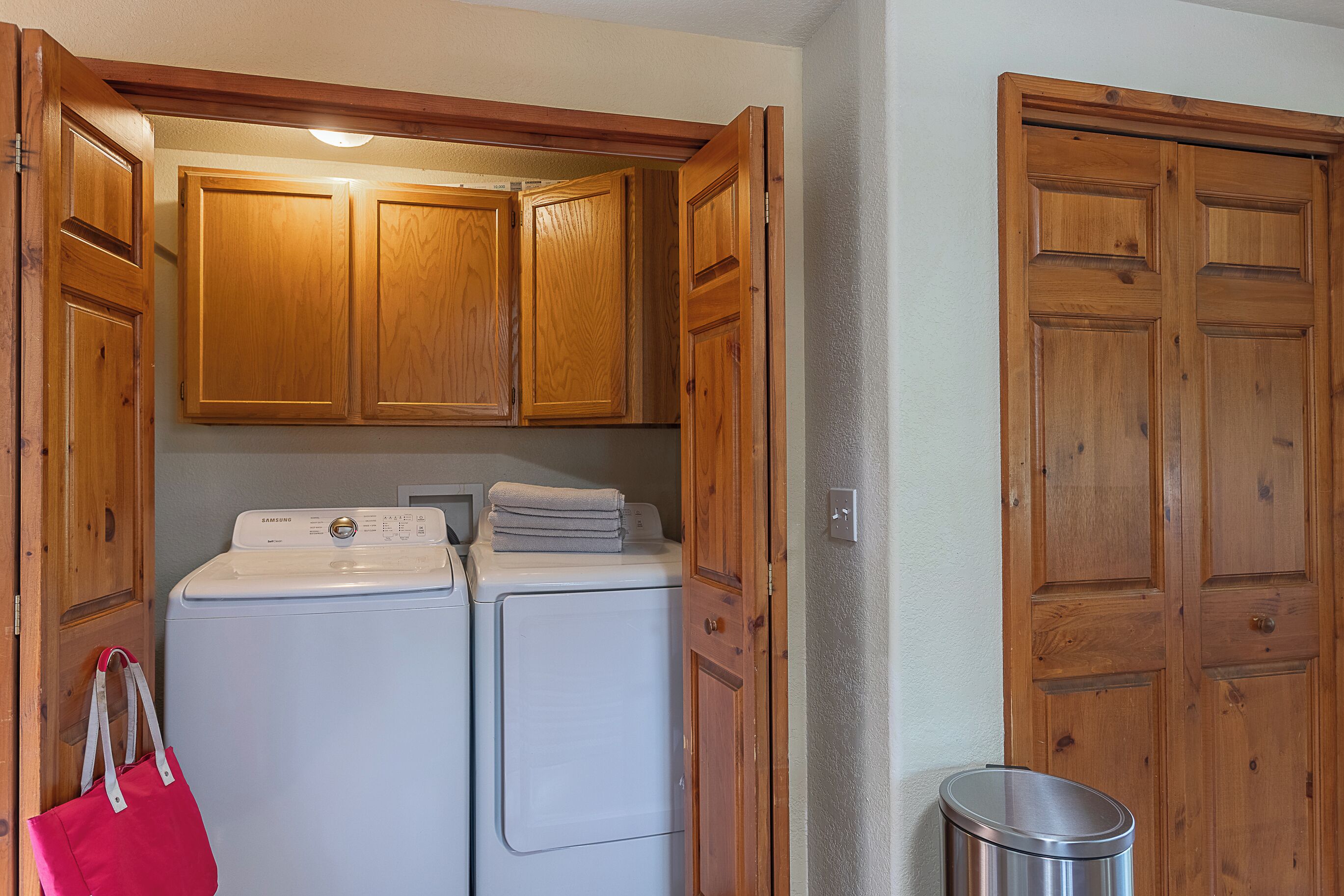 A laundry area with two washing machines, wooden cabinets, and a trash can.