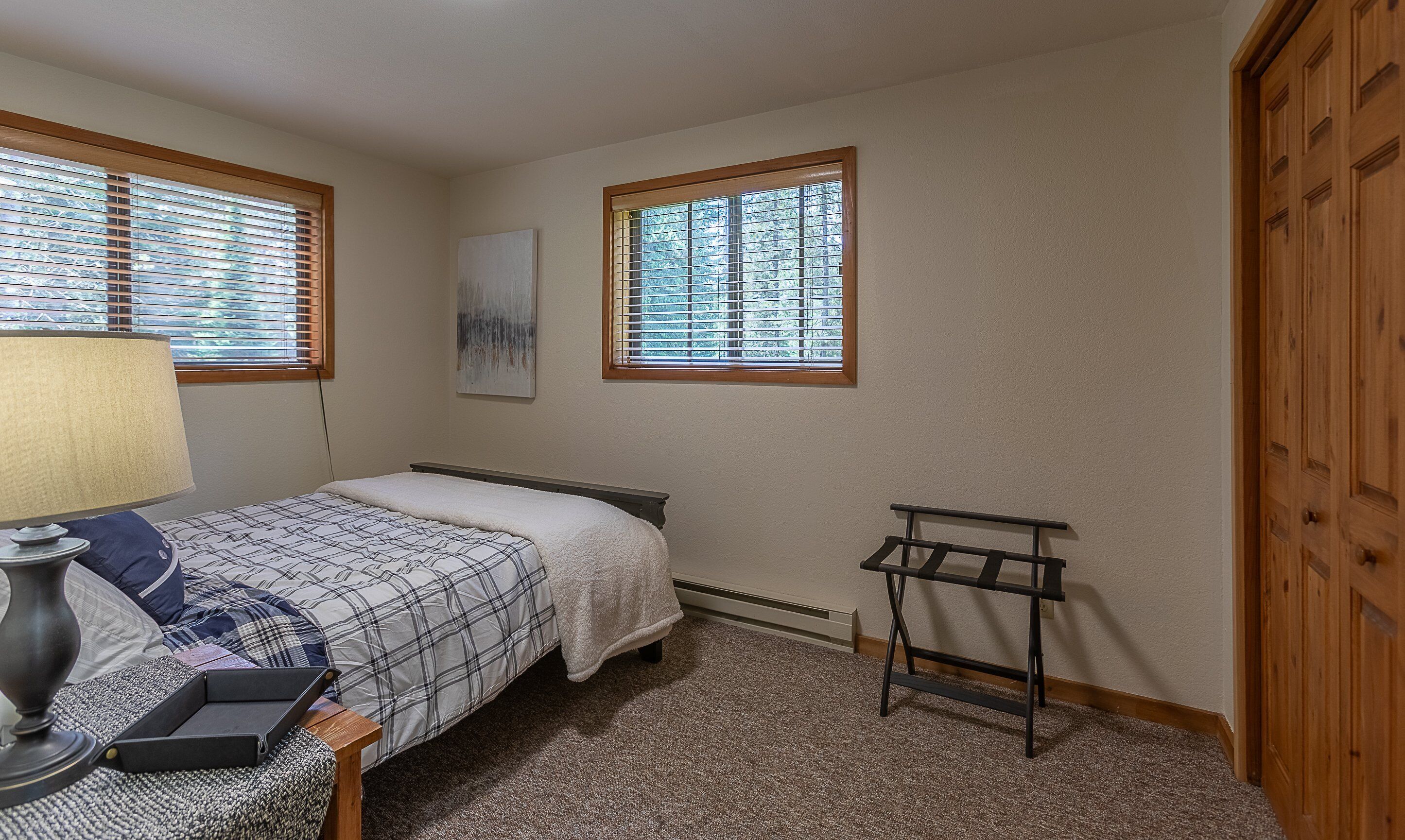 A bedroom with a bed, a bedside table with a lamp, a radiator, a window with blinds, and a wooden wardrobe.