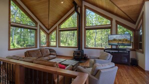 A spacious living room with a wooden ceiling, large windows offering a view of trees, a fireplace, and a television. - Alpine Recharge (Alpine)