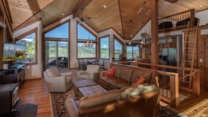 A spacious living area with a high ceiling, large windows, a cozy seating area, and a kitchen in the background. - Alpine Recharge (Alpine)