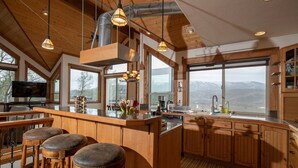 A spacious kitchen with wooden cabinets, a large island with stools, and a view of mountains through the windows. - Alpine Recharge (Alpine)