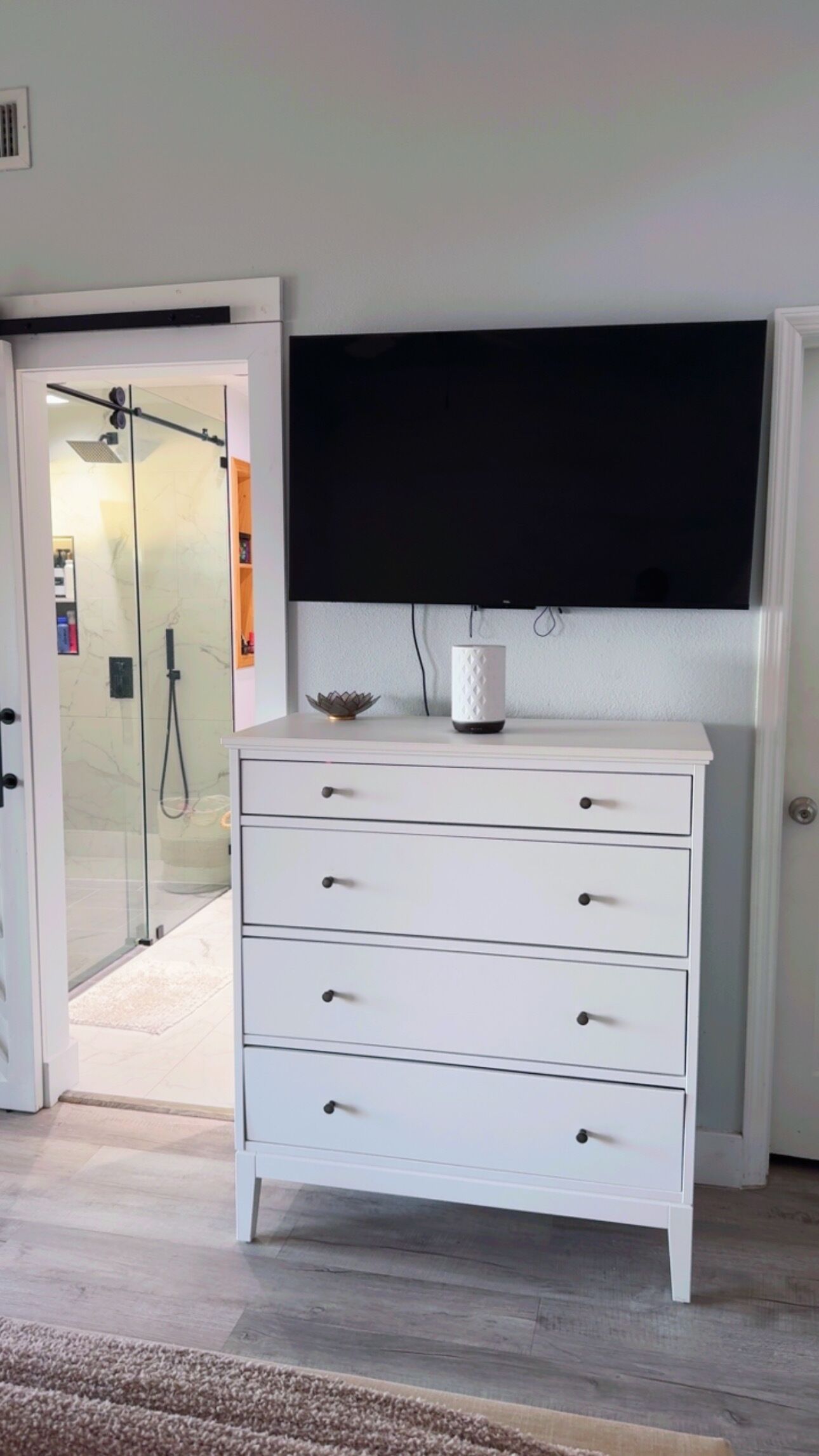 A white dresser with drawers, a flat-screen TV above it, and a glass shower door in the background.
