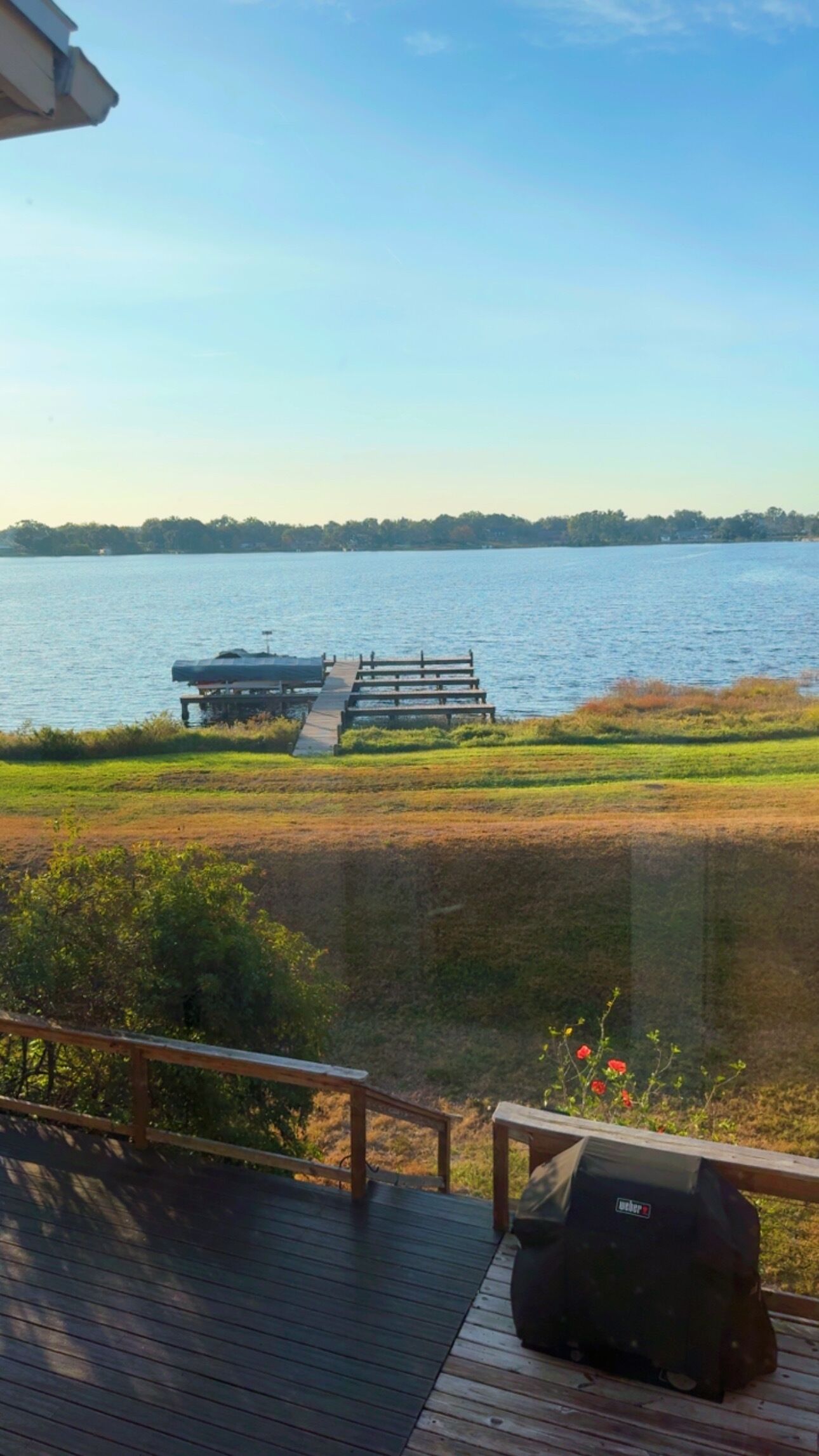 Eine Seeterrasse mit Holzwertung, einem Grill und Blick auf einen Steg, der ins Wasser ragt.
