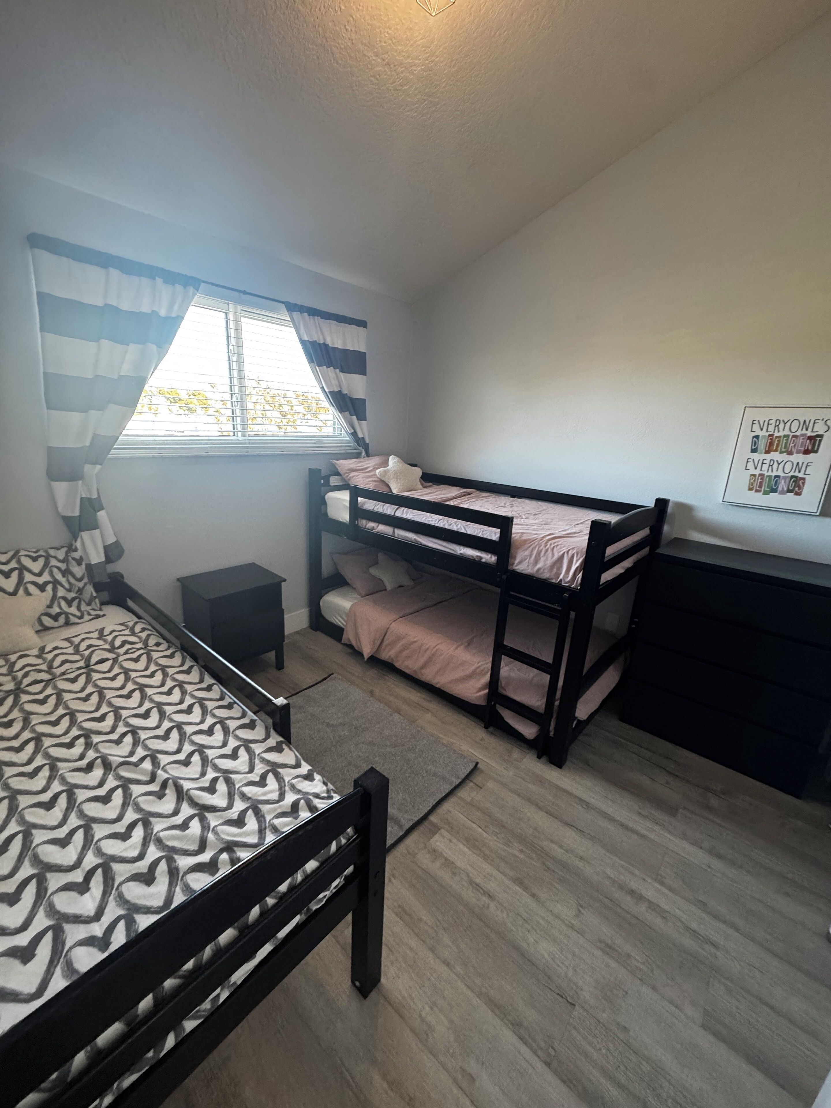 A bedroom with a bunk bed, a single bed, a bedside table, a lamp, a window with curtains, and a motivational poster.