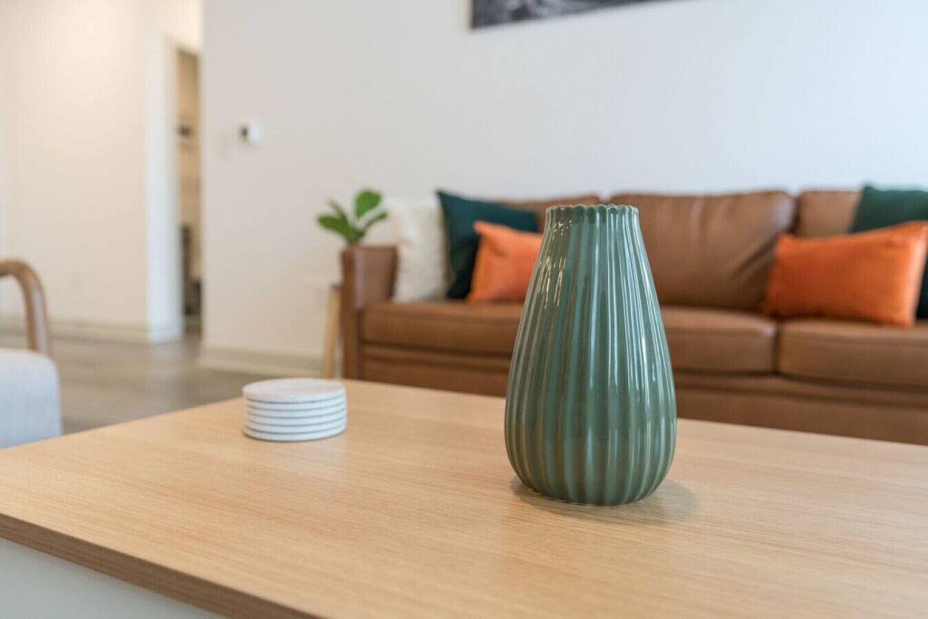 Een houten tafel met een groene vaas en gestapelde borden in een woonkamer met een bruine bank en oranje kussens.