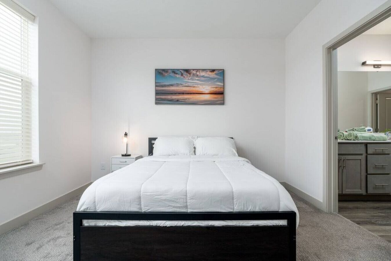 Ein Schlafzimmer mit einem Bett, einem Nachttisch, einem Wandbild und einem durchgehenden Türspalt einsehbaren Badezimmer.