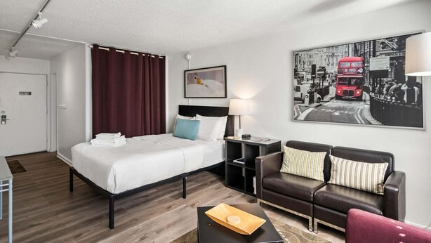 A hotel room with a bed, a desk, a chair, a sofa, a lamp, a picture on the wall, and a door. - Studios On 25th by BCA Furnished Apartments Studio 05 E (Atlanta)