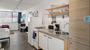 A compact kitchen with a gas stove, microwave, and coffee maker, wooden shelves above the sink, and a window with blinds. - Studios On 25th by BCA Furnished Apartments Studio 05 E (Atlanta)