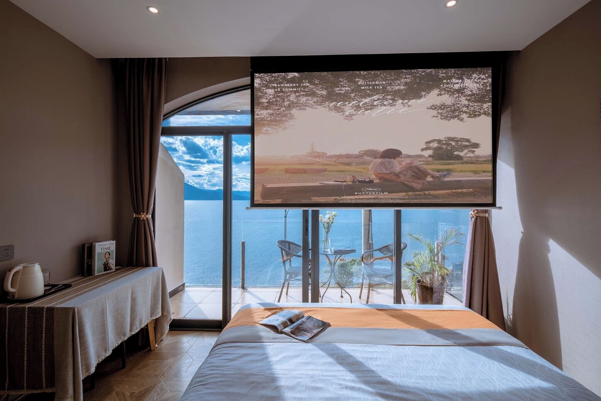 A hotel room with a large window overlooking the sea, a bed with a magazine, a table with a teapot, and a framed picture on the wall.