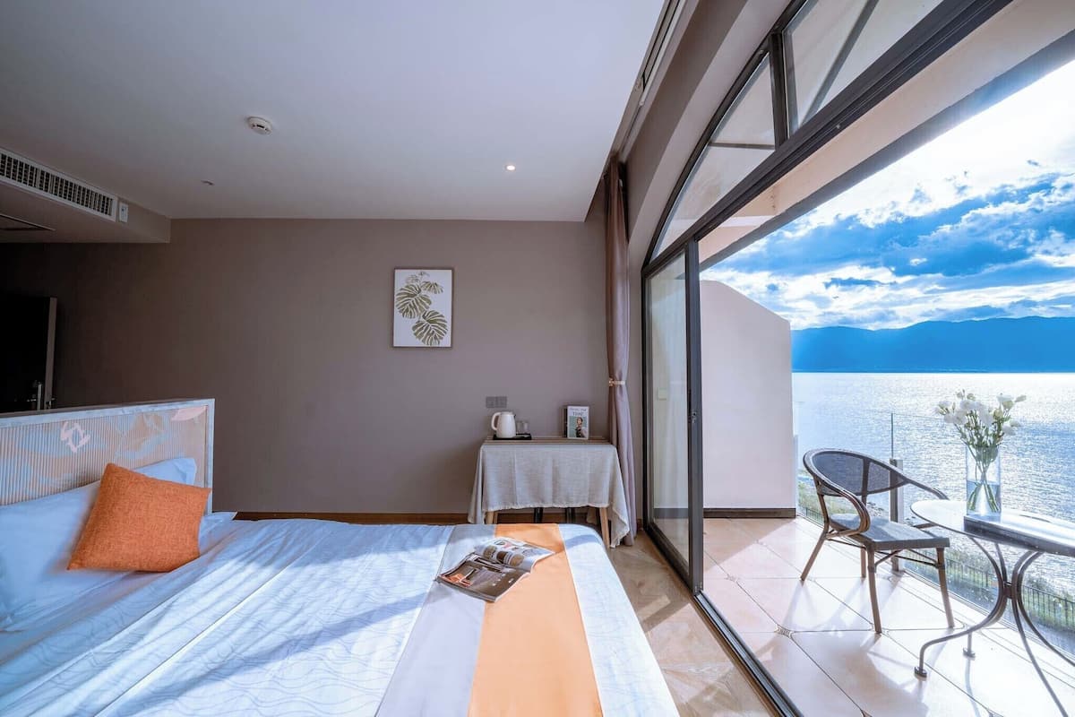 A hotel room with a bed, a small table with a chair, a window with a view of the sea, and a ceiling fan.