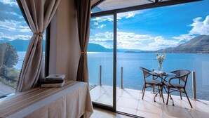 Una habitación con un gran ventanal que da a un lago, una cama, una mesa con sillas y un florero con flores.