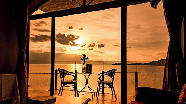 Una habitación con una mesa y sillas frente a un amplio ventanal con vistas a un cuerpo de agua al atardecer.