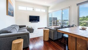 Een moderne woonkamer met een grijze modulaire bank, een houten salontafel en een eethoek met een houten tafel en stoelen. Een flatscreen televisie is aan de muur bevestigd, en er zijn grote ramen met uitzicht op de stad.