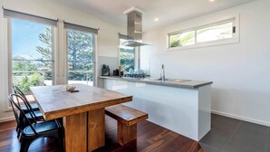 Een moderne keuken met een houten eettafel en een bank, grote ramen en uitzicht op groen.