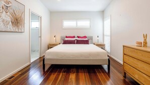 Een moderne slaapkamer met een groot bed, houten vloeren, een nachtkastje met lamp, een dressoir en een raam met blinds.