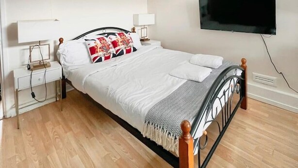 Un lit bien fait, avec une literie blanche et des coussins décoratifs, une table de chevet avec une lampe et un téléviseur à écran plat fixé au mur.