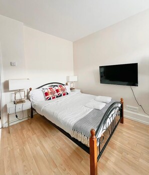 Un lit bien fait, avec une literie blanche et des coussins décoratifs, une table de chevet avec une lampe et un téléviseur à écran plat fixé au mur.