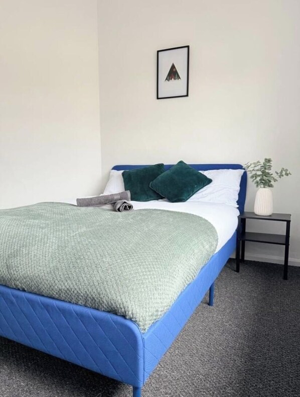 A bed with a blue headboard, a green blanket, and a pillow. There is a small white vase with a plant on a nightstand beside the bed. - 3 bedroom house, Brisco Terrace, Egremont (Egremont)