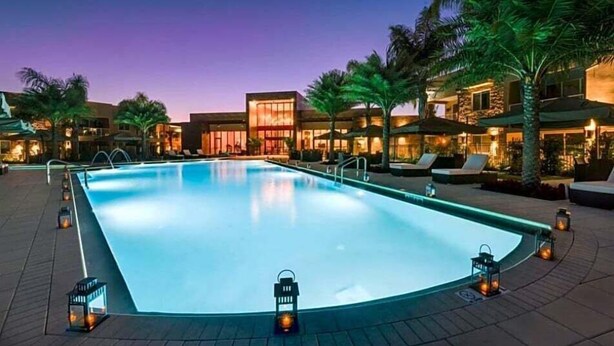 Una piscina con sillas de descanso y palmeras, rodeada de edificios con iluminación cálida.