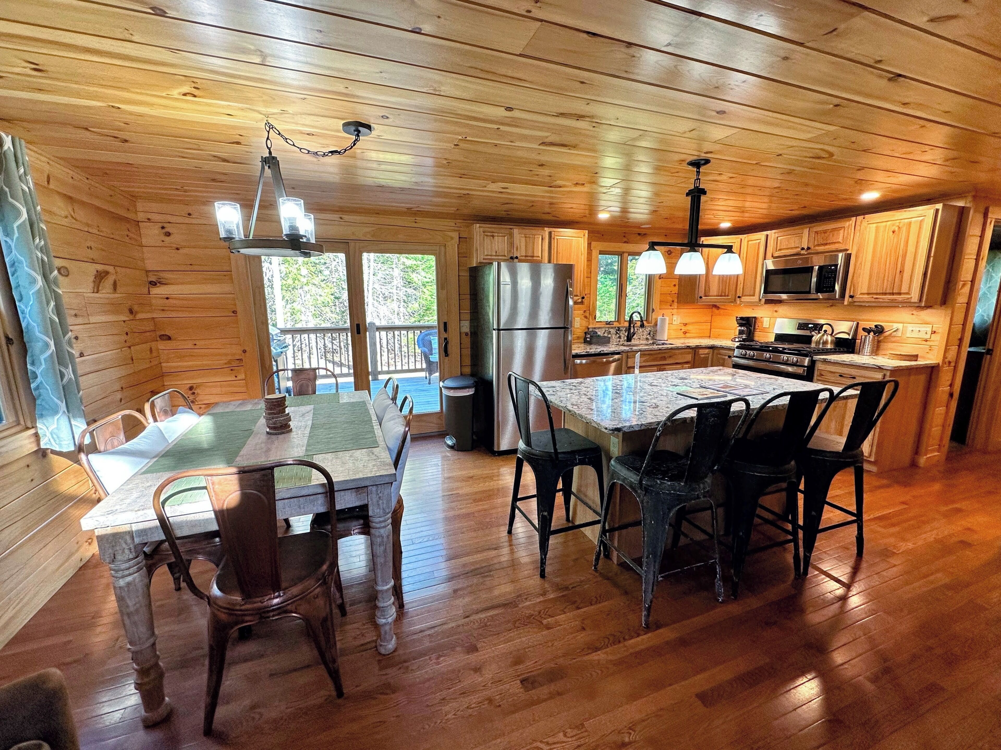 A wooden cabin interior with a dining area, kitchen, and a view of the outdoors.