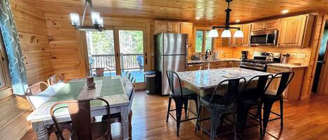 A wooden cabin interior with a dining area, kitchen, and a view of the outdoors.