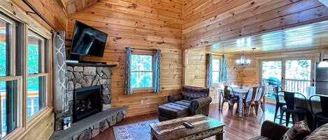 A cozy cabin interior with a stone fireplace, wooden walls, a television mounted above it, and a dining area with a table and chairs.