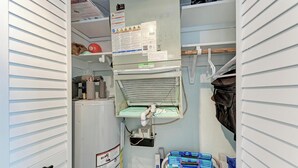 Un cuarto de servicio compacto con calentador de agua, panel de control con indicaciones de seguridad y un pequeño espacio de almacenamiento con bolsas y una maleta azul.