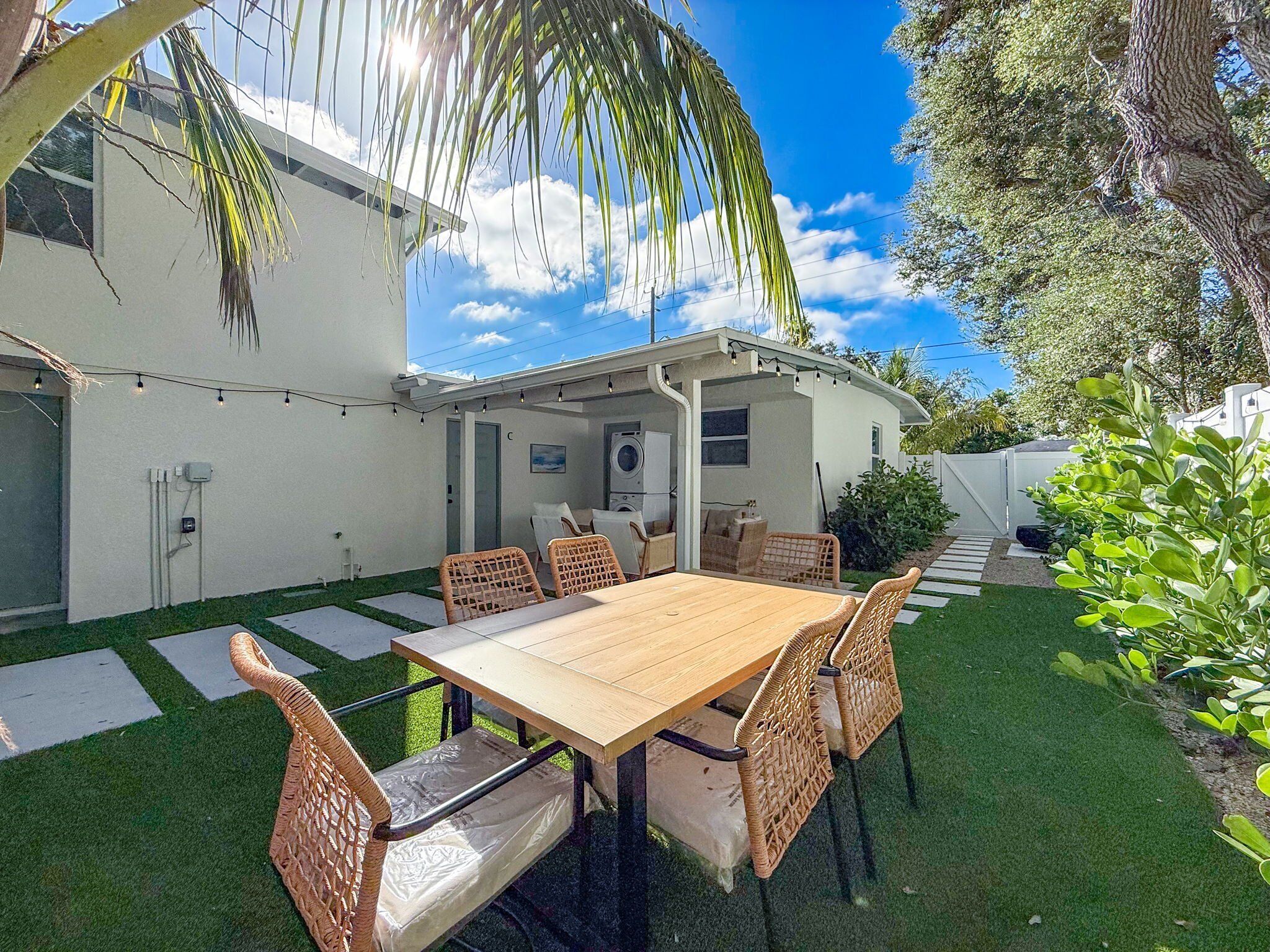 Un patio con una mesa de madera y sillas de mimbre, rodeado de un pequeño césped y un edificio blanco con una zona cubierta.