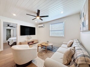 A modern living room with a flat-screen TV, ceiling fan, and a large window with blinds. - 3716 7th Ave W (Bradenton)