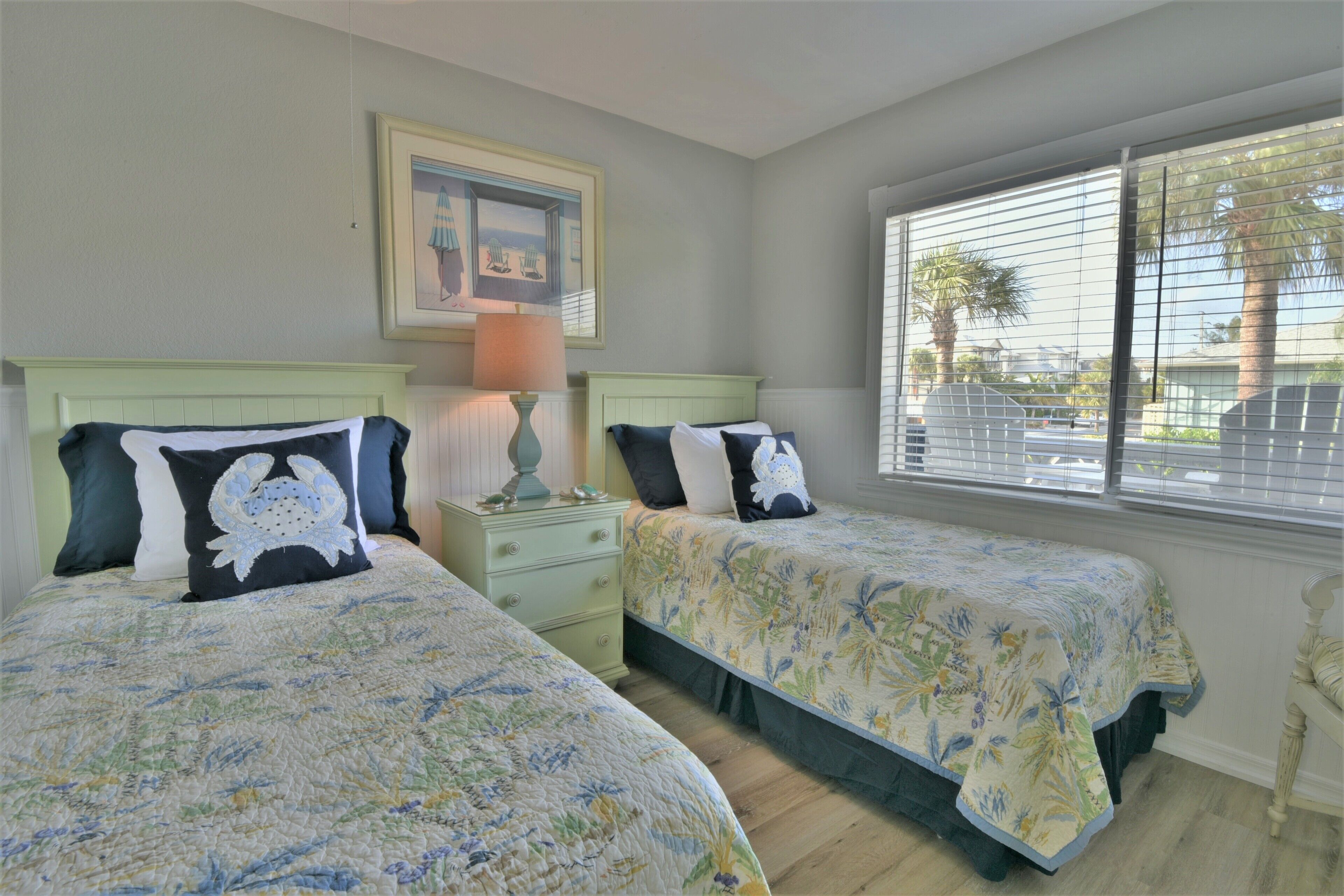 A bedroom with two beds, each with floral bedspreads, a nightstand with a lamp, a framed picture on the wall, and a window with blinds.