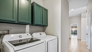 A laundry room with two white front-loading washers and dryers, green cabinets above, and a hallway leading to a door. - Relaxing 4BR Retreat w/ Refreshing Pool (Indio)