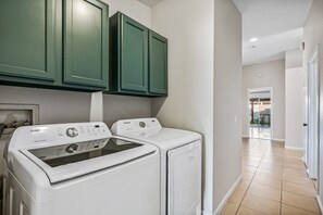 A laundry room with two white front-loading washers and dryers, green cabinets above, and a hallway leading to a door. - Relaxing 4BR Retreat w/ Refreshing Pool (Indio)